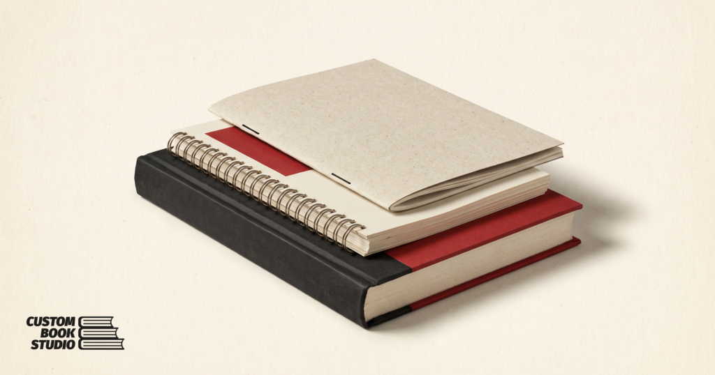 Custom Book Studio — a small stack of finished printed books of mixed bindings — a coil workbook, a booklet and a red hardcover (watermarked).